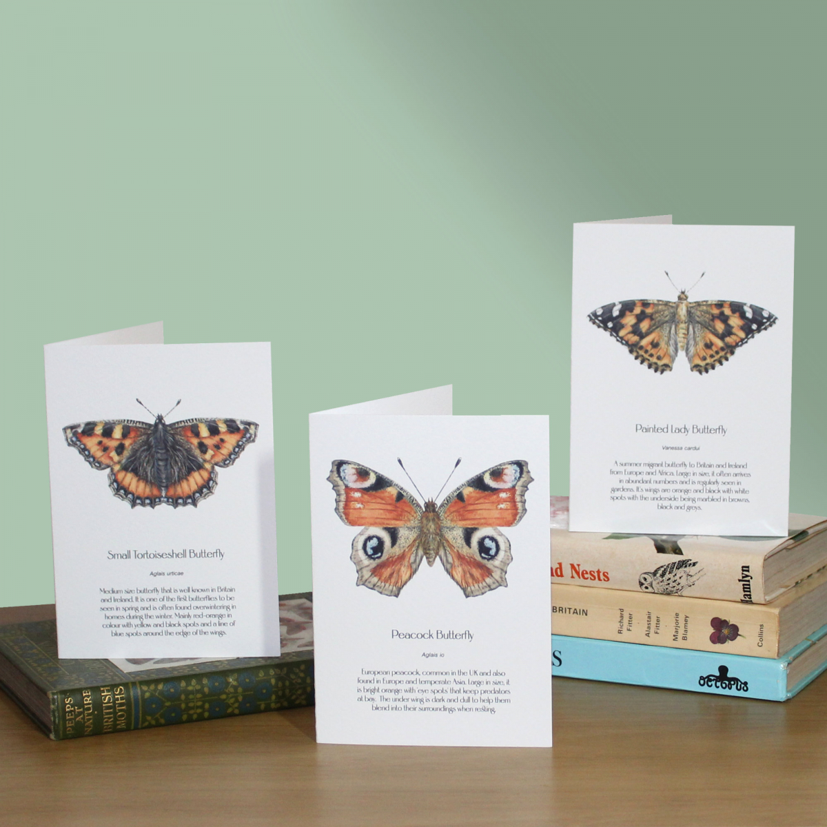 3 greetings cards with butterflies and information about them. They are sitting on a pile of books on a green background.